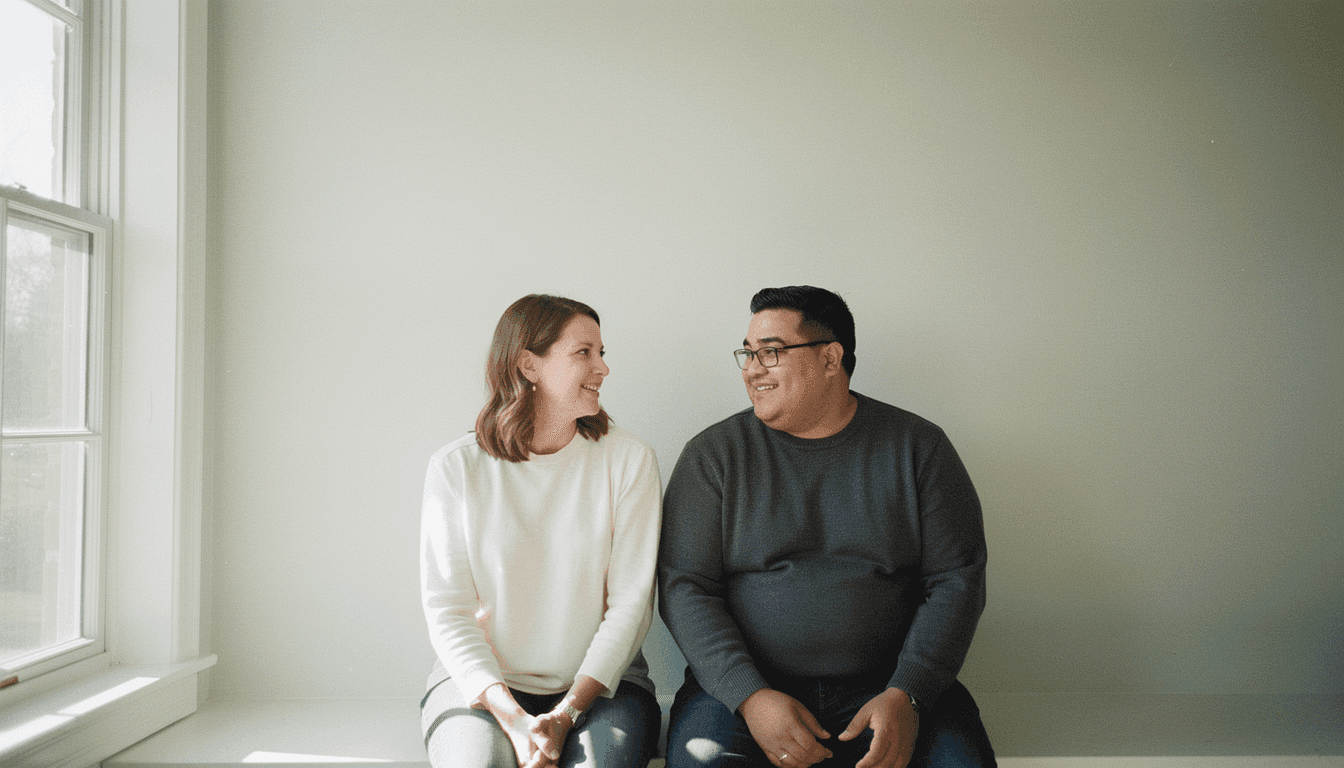 A couple sits together on a bright windowsill, talking, one holding a purple mug.