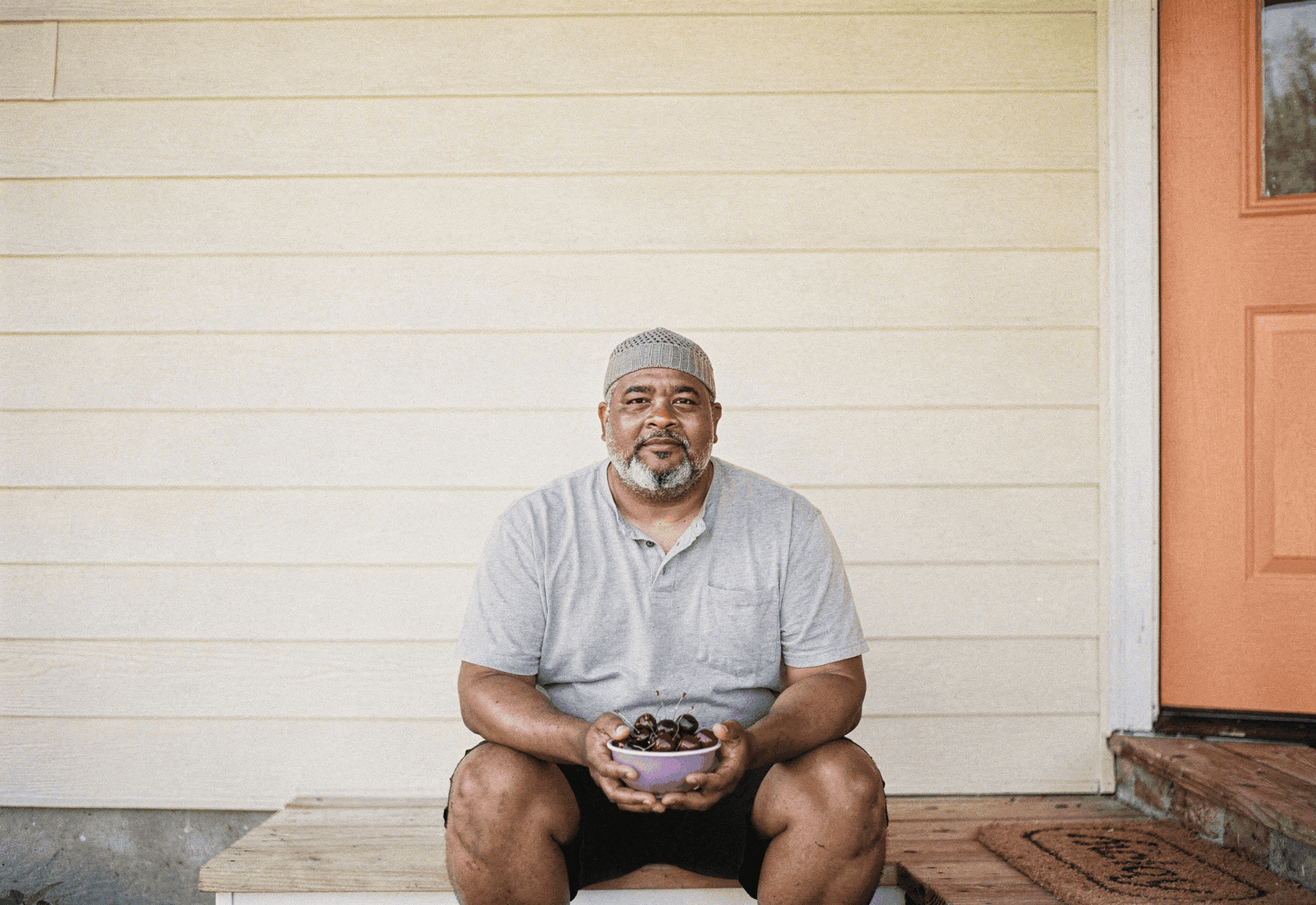 A man in his early sixties sits on his front porch step with a bowl of cherries in late-afternoon sun.