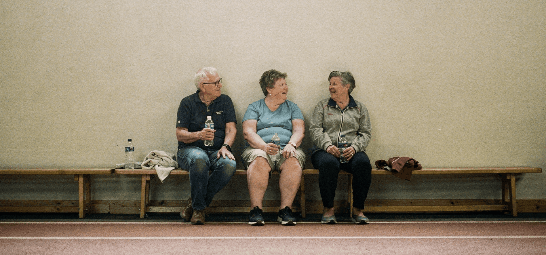 Three neighbors rest on a bench beside an indoor walking track, holding water bottles and chatting.