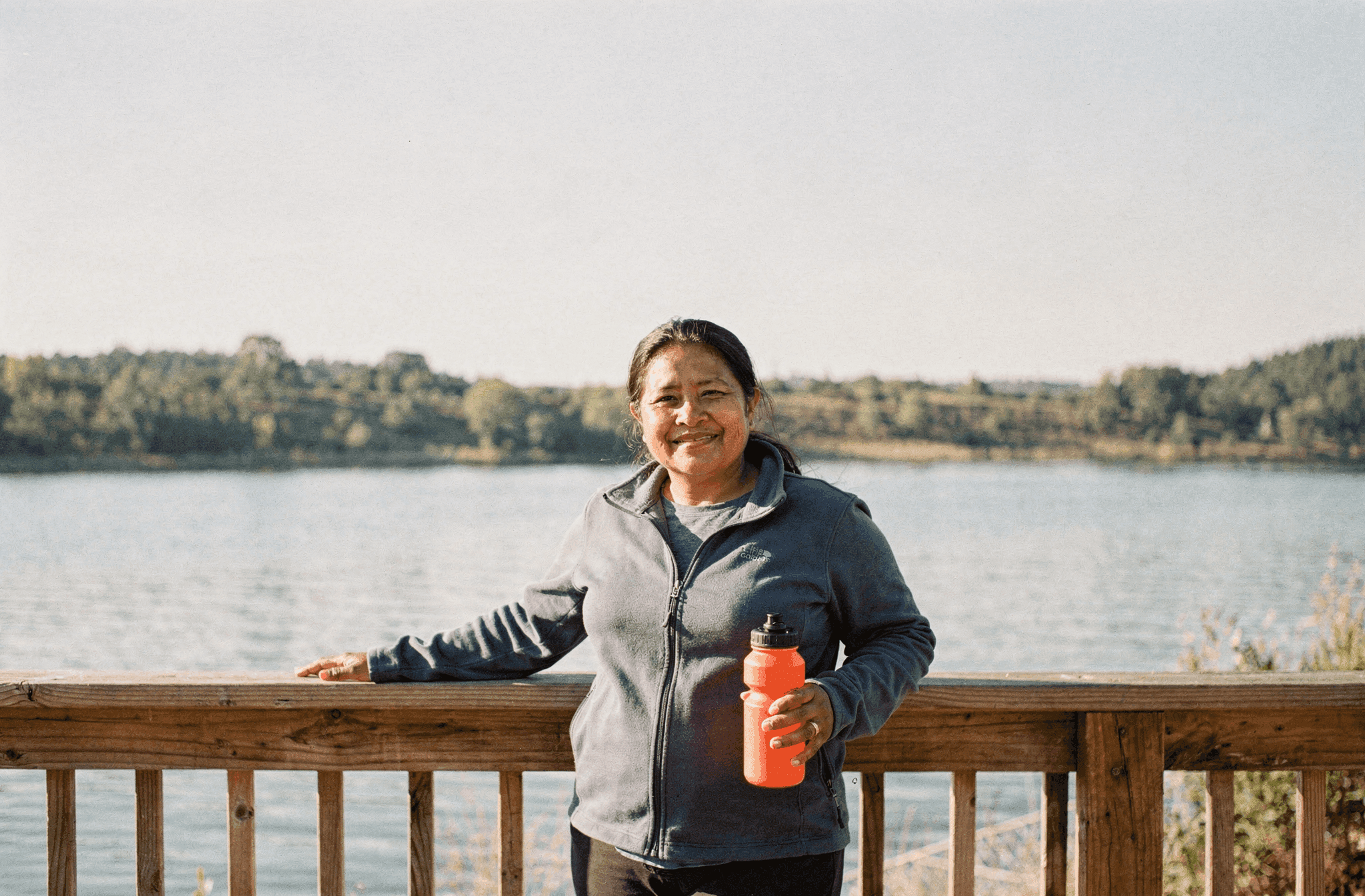 A woman in her late thirties stands at a lake overlook on a clear morning holding a water bottle.